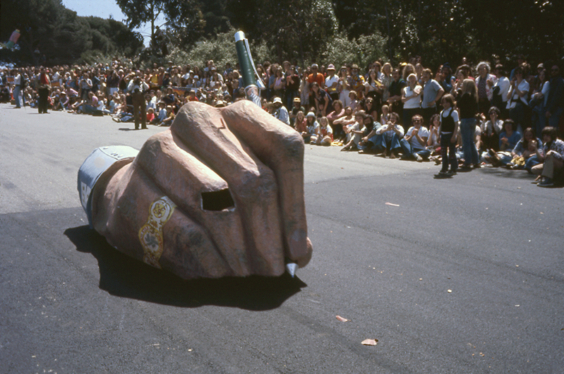 What looks like a giant hand holding a pen rolls down a hill. Spectators stand in the background